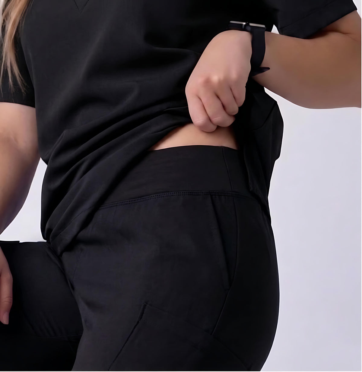 A close-up of a person wearing black athletic scrub pants and a matching black top, lifting the hem of the top to show the fit and waistband.