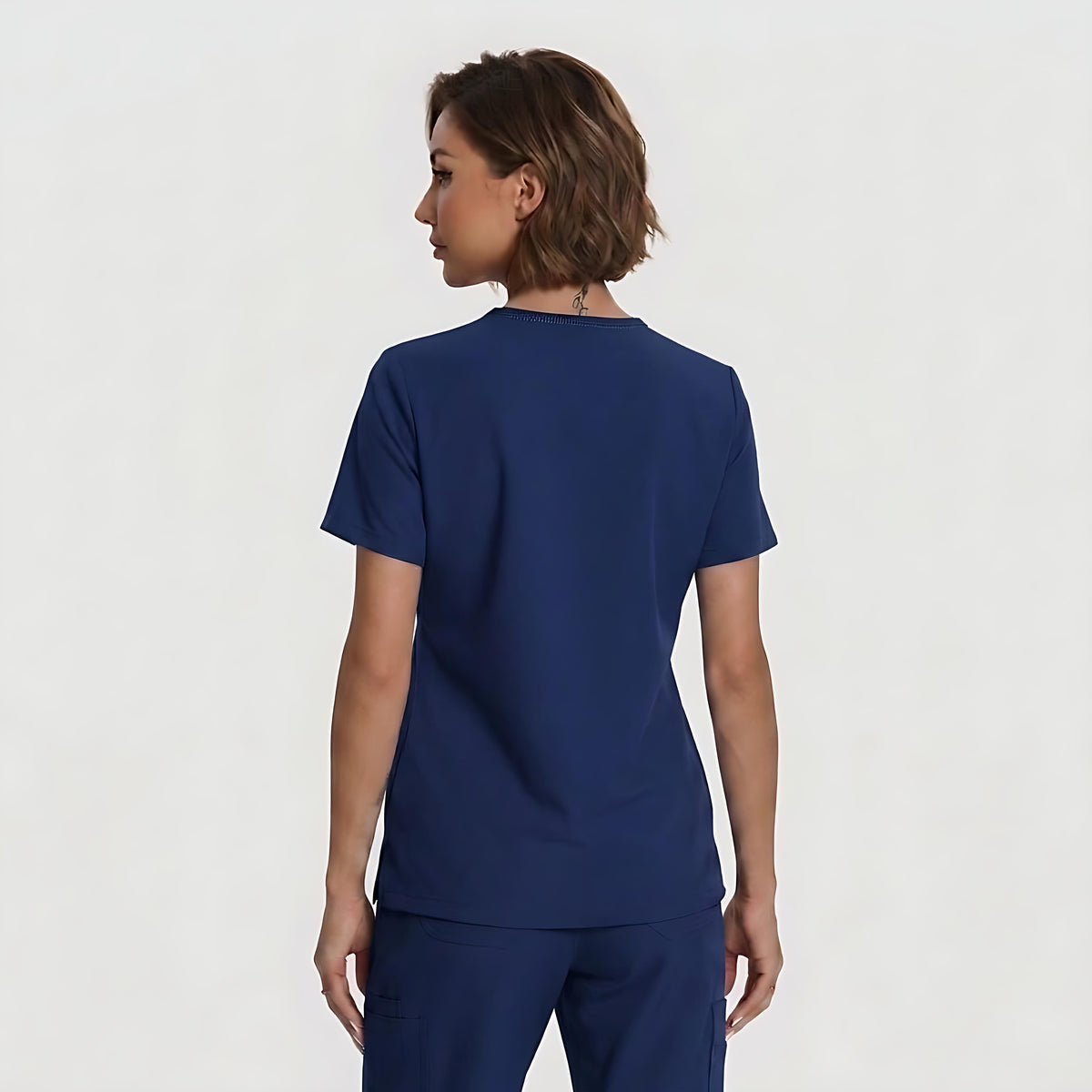 A woman with short brown hair is shown from the back wearing a navy blue scrub top and matching pants against a light gray studio background.