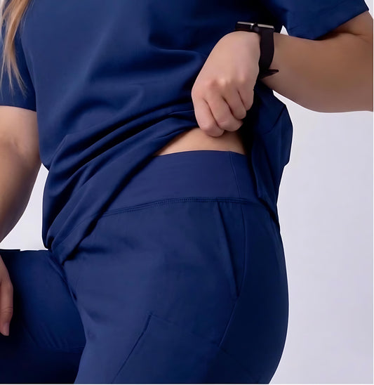 A close-up of a person wearing navy blue scrub-style clothing, lifting the top to show the back waistband and side pocket area, against a light gray studio background.