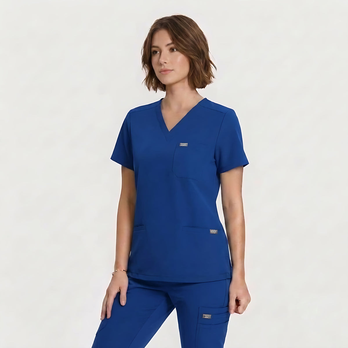 A woman modeling a royal blue scrub top and matching scrub pants against a clean, light off-white studio background.