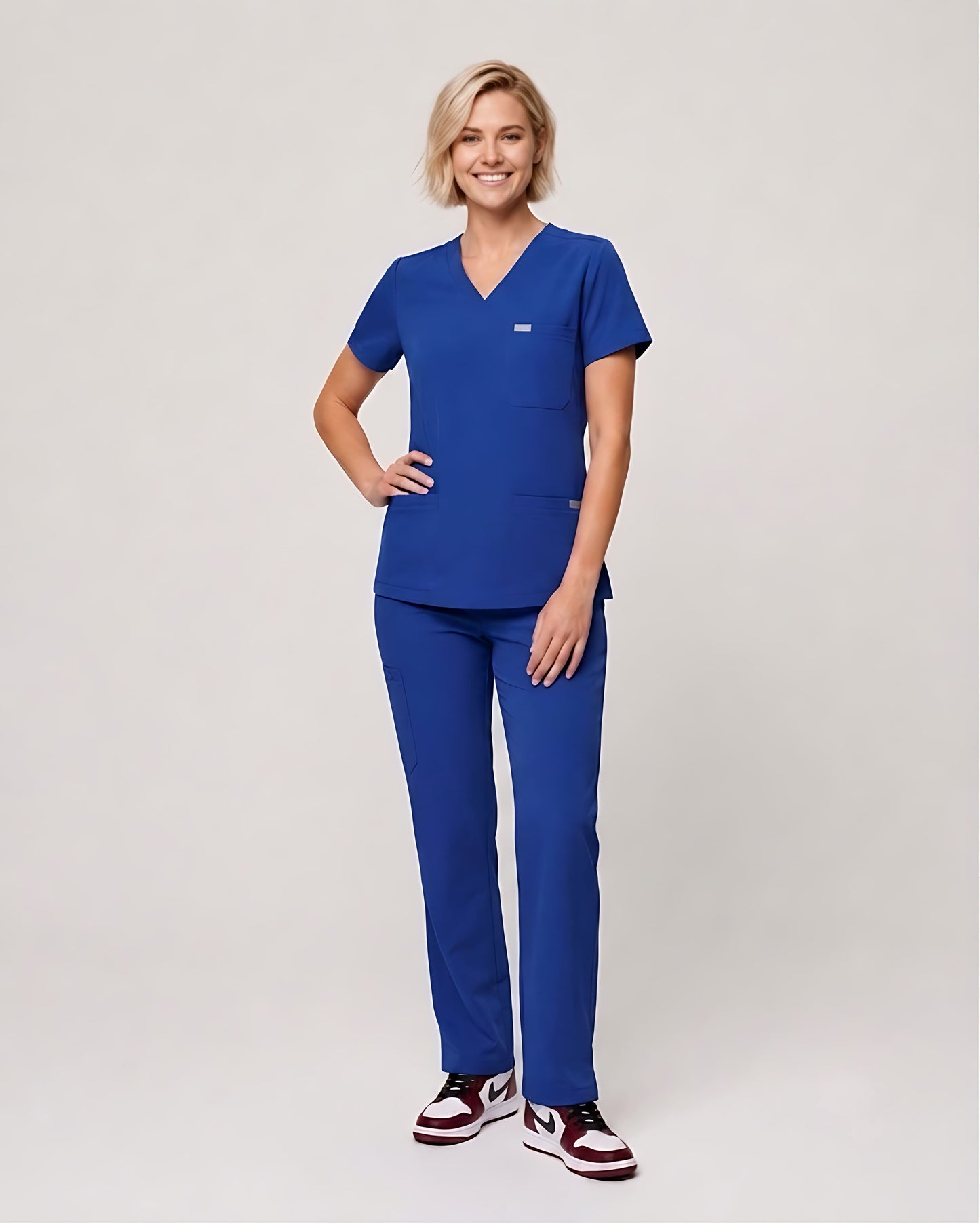 A woman modeling a matching royal blue scrub set, consisting of a short-sleeve V-neck top and straight-leg pants, paired with maroon and white sneakers, against a light gray studio background.