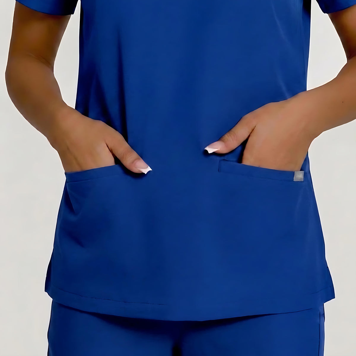 A close-up of a person wearing a royal blue scrub top with two front pockets, with both hands resting in the pockets. The image is cropped from the chest down, showing the fit and pocket details against a light gray background.