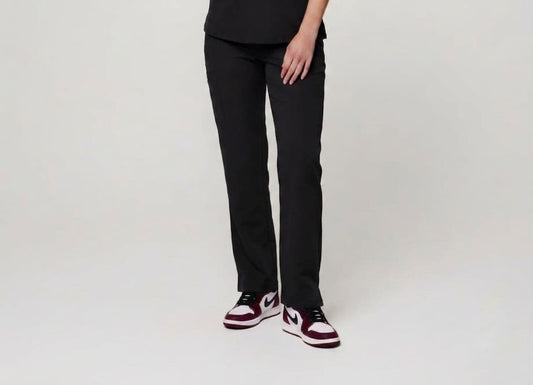 A person wearing black straight-leg pants and black, white, and burgundy sneakers, standing against a plain light gray studio background.