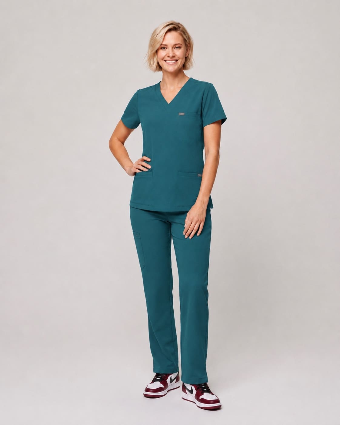 A woman modeling a matching teal scrub set consisting of a V-neck short-sleeve top and straight-leg pants, paired with maroon and white sneakers, against a light gray studio background.