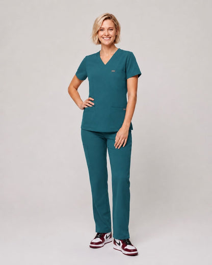 A woman modeling a matching teal scrub set consisting of a V-neck short-sleeve top and straight-leg pants, paired with maroon and white sneakers, against a light gray studio background.