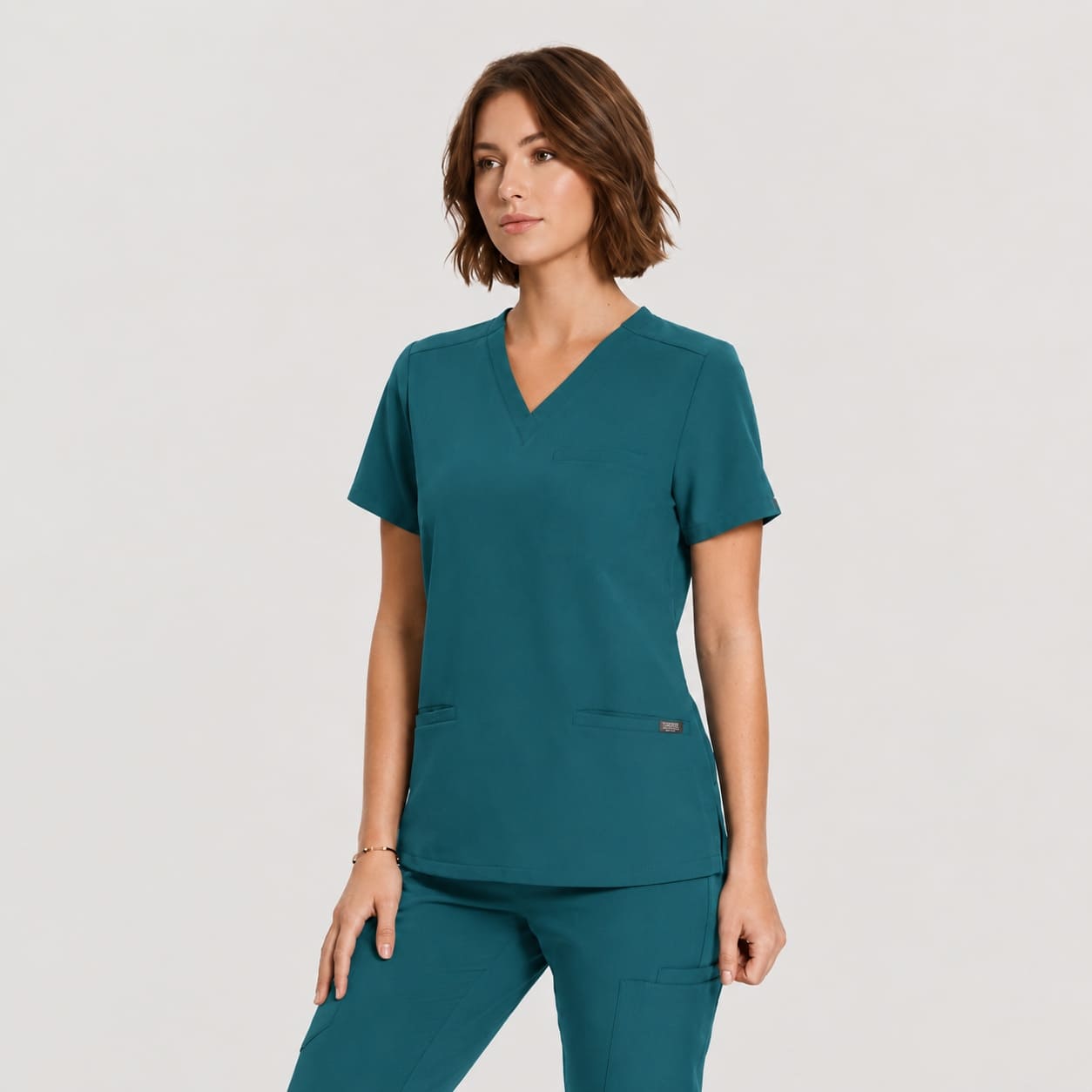 A woman modeling a teal V-neck scrub top and matching scrub pants against a light gray studio background.