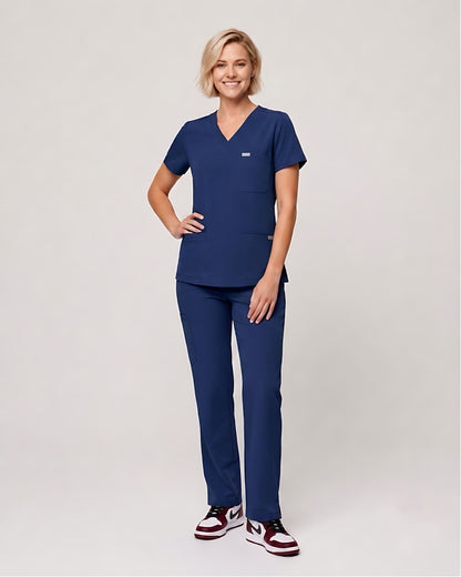 A woman modeling a navy blue scrub set consisting of a short-sleeve V-neck top and matching straight-leg pants, paired with maroon and white sneakers, against a light gray studio background.