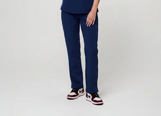 A person wearing navy blue scrub pants with a matching navy top and white, burgundy, and black sneakers, shown from the waist down against a light gray studio background.