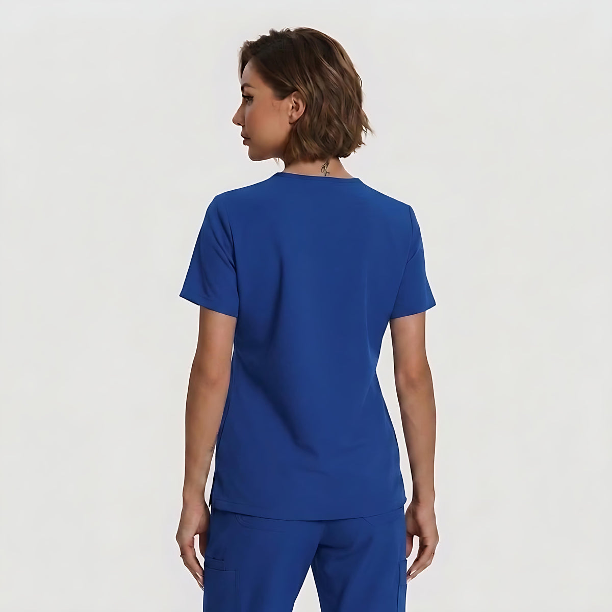 A woman shown from the back wearing a royal blue short-sleeve scrub top and matching scrub pants against a plain light gray studio background.