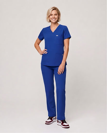 A woman modeling a matching royal blue scrub set, consisting of a short-sleeve V-neck top and straight-leg pants, paired with maroon and white sneakers, against a light gray studio background.