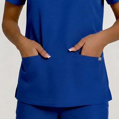 A close-up of a person wearing a royal blue scrub top with two front pockets, with both hands resting in the pockets. The image is cropped from the chest down, showing the fit and pocket details against a light gray background.