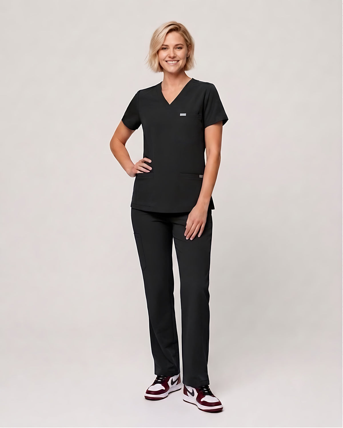 A woman modeling a black V-neck scrub top and matching black scrub pants against a light gray studio background.