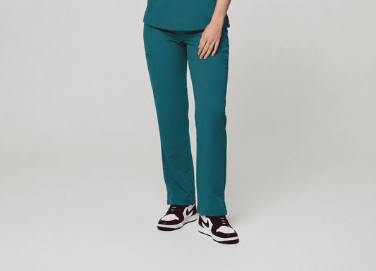 A person wearing teal scrub pants and a matching teal top, standing against a light gray studio background. The photo is cropped from the waist down, showing the pants, hand, and black-and-white sneakers.