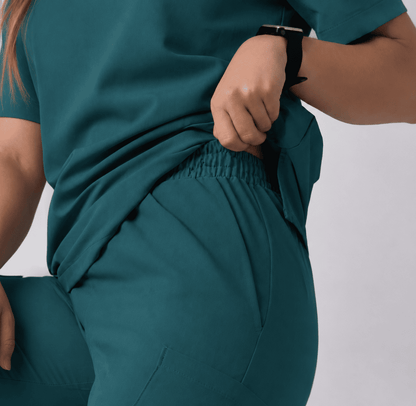 A close-up of a person wearing teal scrub-style clothing, pulling the top slightly away to show an elastic waistband and side pocket detail against a light gray background.